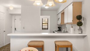 A modern kitchen with a white countertop, wooden cabinets, and a built-in sink. There are two wooden stools and a small potted plant on the counter.