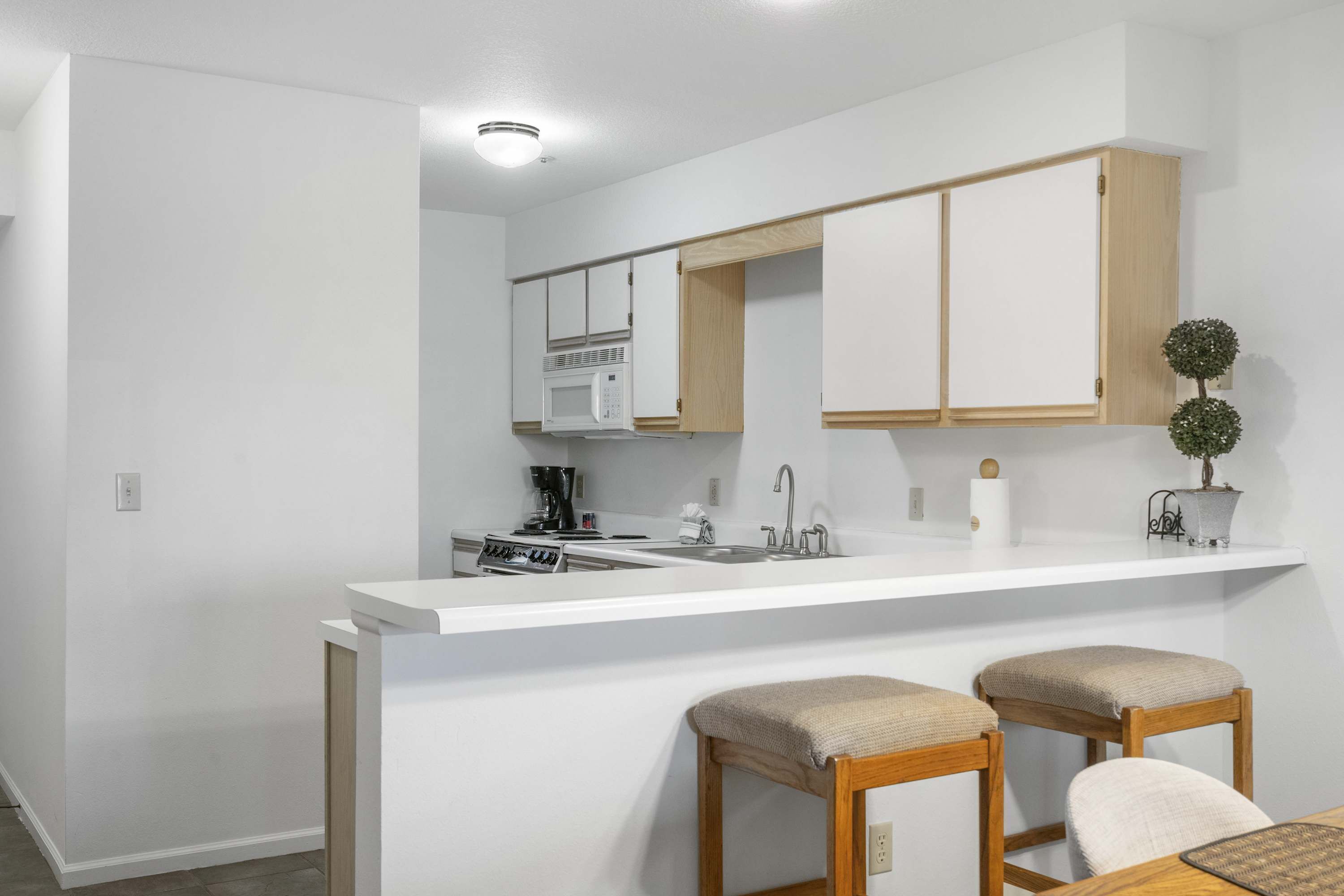 Une cuisine moderne avec des meubles blancs, un four à micro-ondes et un évier. On y trouve deux tabourets en bois avec des coussins.