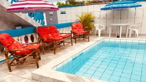En swimmingpool med kakelbeklädda kanter, omgiven av flätade loungestolar med röda kuddar, ett vitt bord och blårandiga parasoller.
