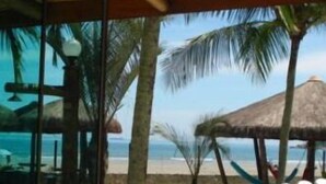 Vistes a la platja des d'una habitació amb paret de vidre, incloent una construcció amb teulada de palla i gandules.