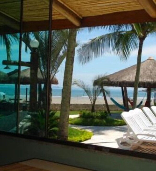 A beach view from a room with a glass wall, including a thatched-roof structure and lounge chairs. - Valentina Praia Hotel (São Sebastião)