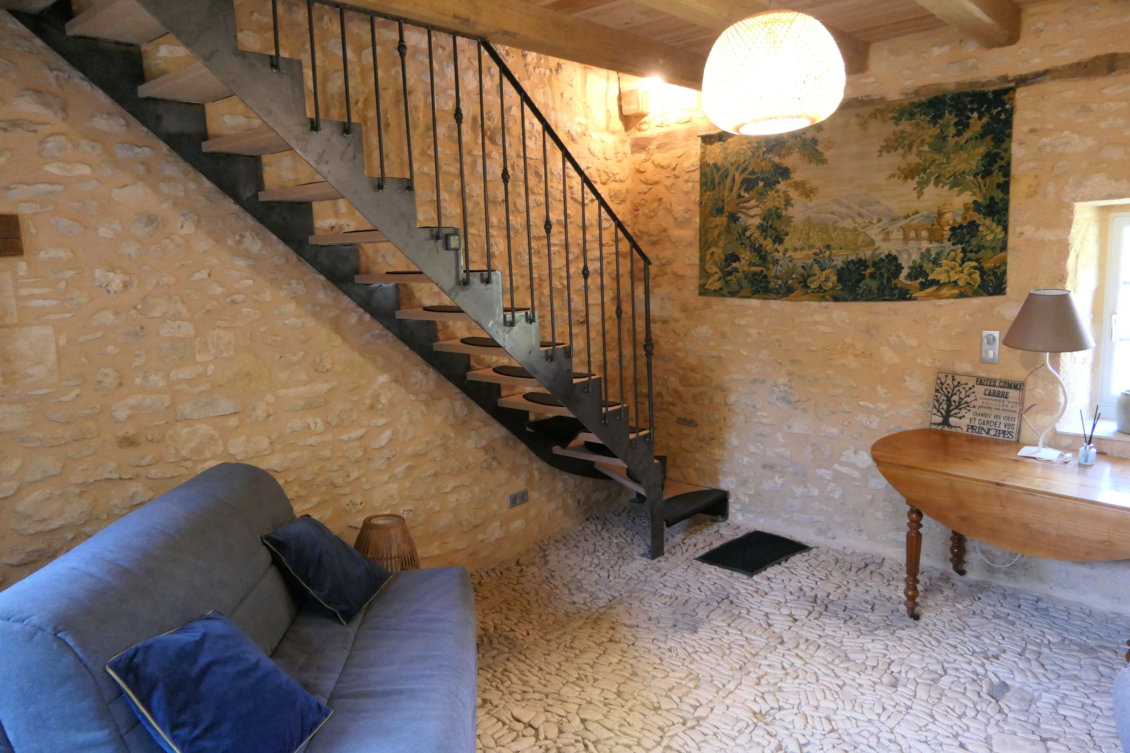 Una habitación con paredes de piedra, una escalera de madera, un sofá con cojines, una mesa de madera y una lámpara de pared.