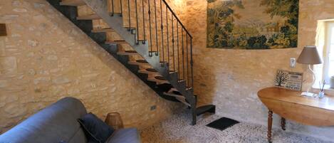 Una habitación con paredes de piedra, una escalera de madera, un sofá con cojines, una mesa de madera y una lámpara de pared.