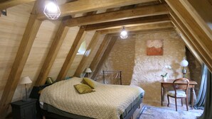 A cozy attic bedroom with a wooden beam ceiling, a bed with a quilt, a desk with a chair, and a potted plant.