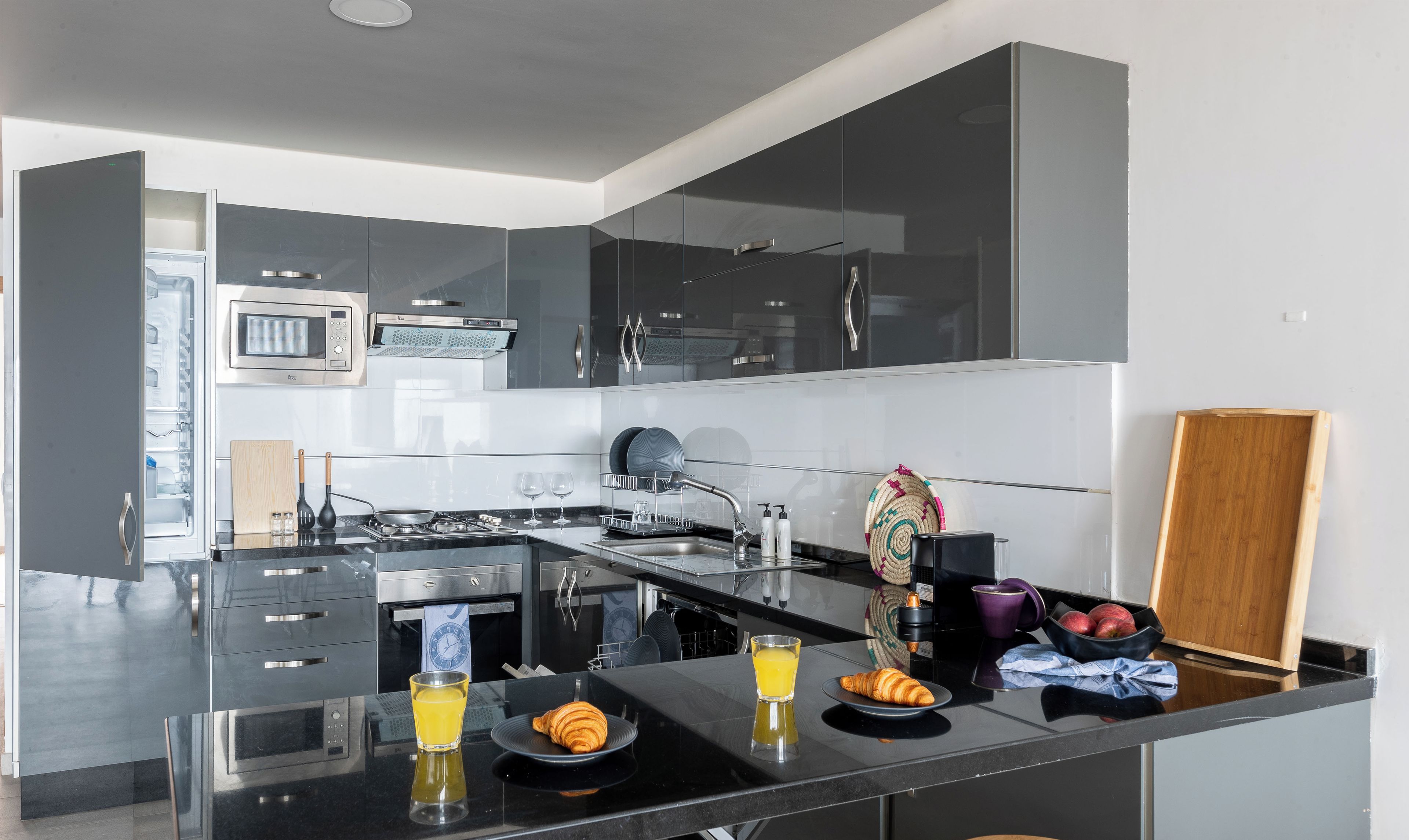 Een moderne keuken met een ingebouwde oven, magnetron en kookplaat. Er is een ontbijt klaargezet met croissants, sap en een fruitschaal.