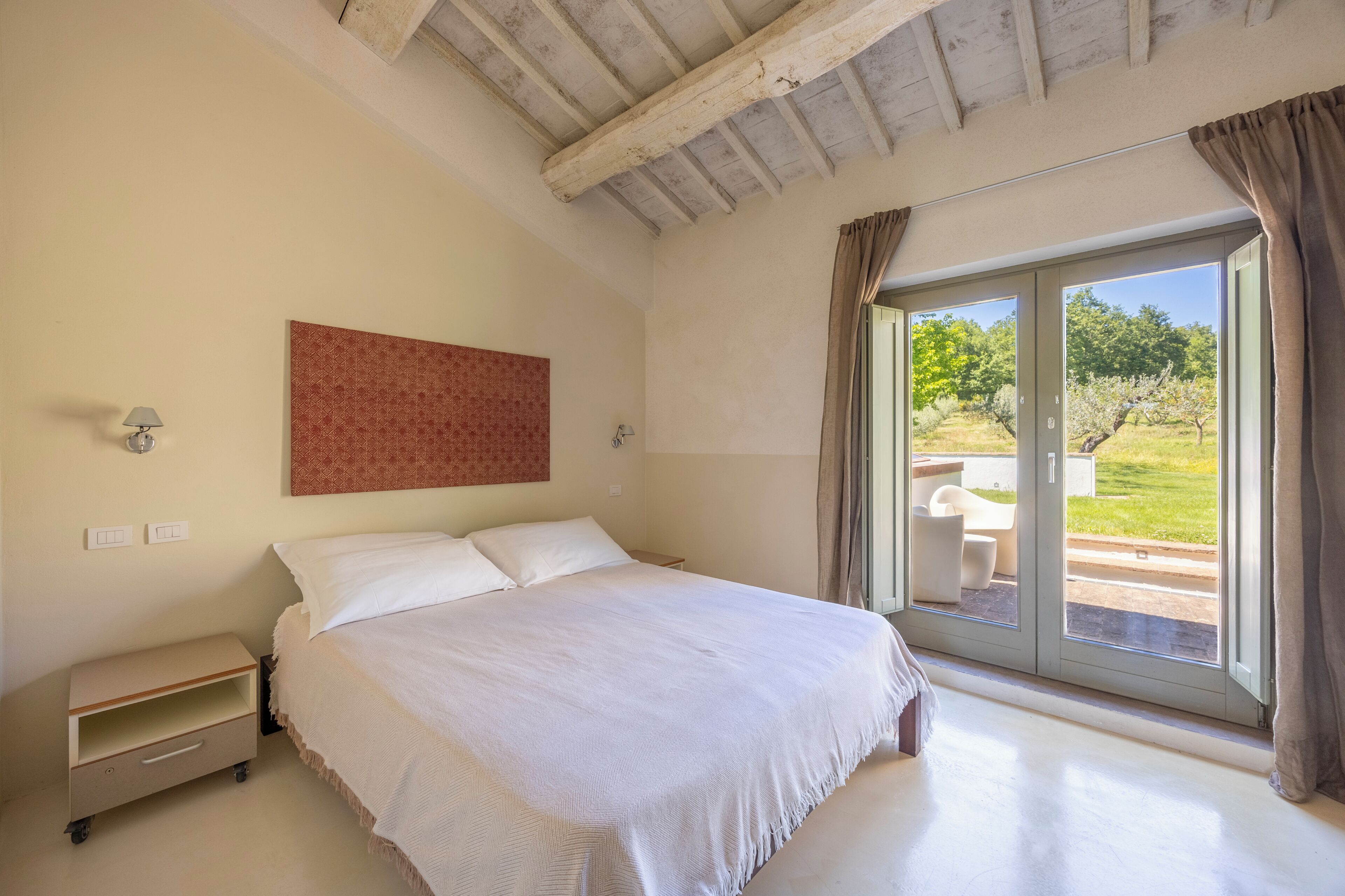 A bedroom with a bed, a nightstand, a wall-mounted lamp, a sliding glass door to an outdoor area, and a red patterned wall.