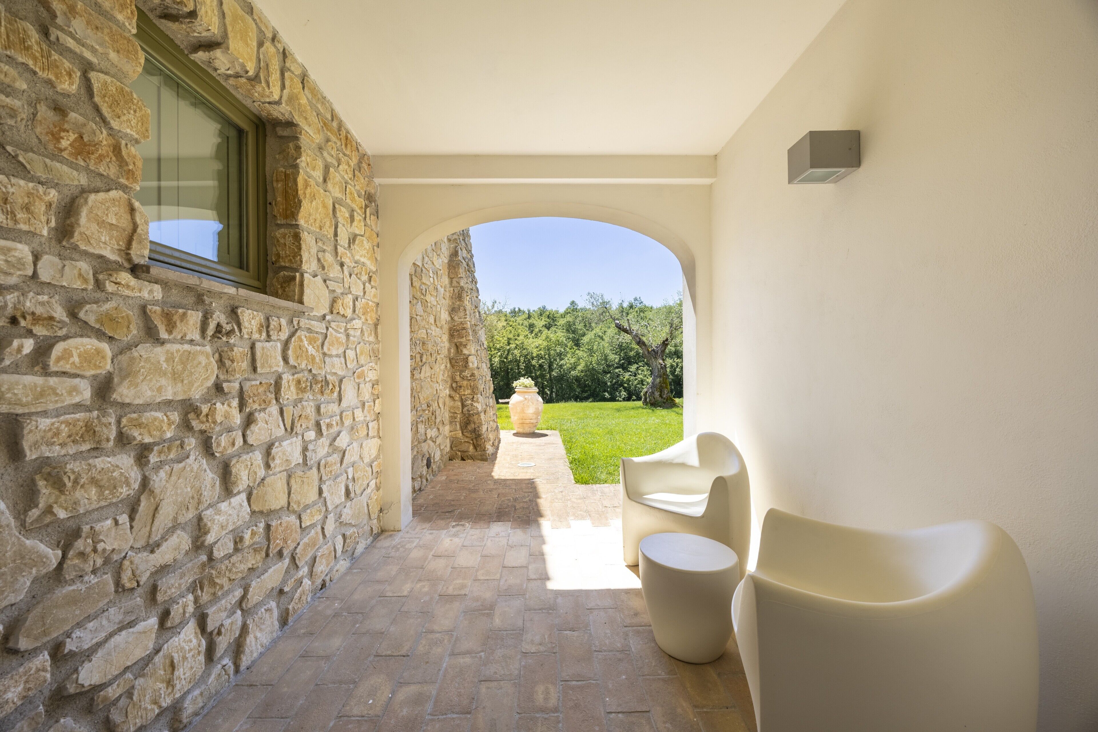 A stone-walled corridor with a view to a garden, a window, and a seating area with a table.