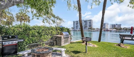 A patio with a grill, fire pit, and seating area overlooking a body of water and cityscape.