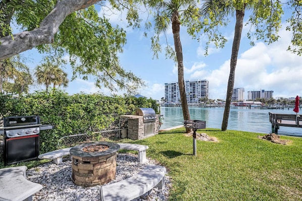 A patio with a grill, fire pit, and seating area overlooking a body of water and cityscape.