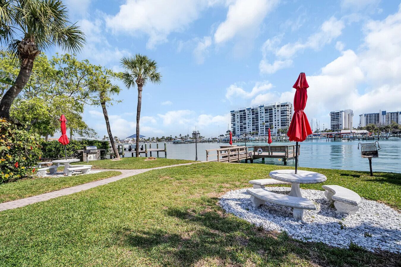 Une terrasse au bord de l’eau, agrémentée de parasols rouges, de bancs blancs et offrant une vue dégagée sur le port de plaisance et les bâtiments qui se dressent à l’arrière-plan.
