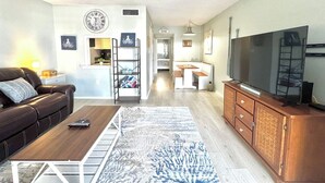 Una sala moderna con sofá, mesa de centro y mueble para televisión. Se ve una cocina al fondo, con mesa y sillas.