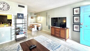 Una sala de estar con una mesa de centro de madera, un televisor sobre un mueble de madera, una zona de comedor con un banco, y una cocina con una encimera amarilla.