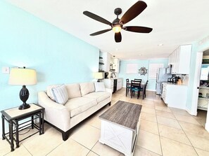 Sala de estar con sofá, mesa de centro y ventilador de techo. Incluye una zona de cocina con alacenas y mesa de comedor.