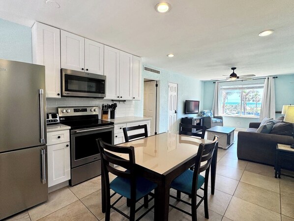 A modern kitchen with stainless steel appliances, a dining area with a glass table, and a living room with a sofa and TV.