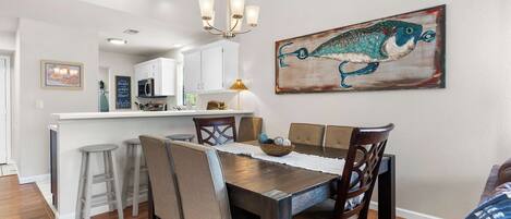A dining area with a wooden table, chairs, and a chandelier. There is a kitchen in the background with white cabinets and a microwave.