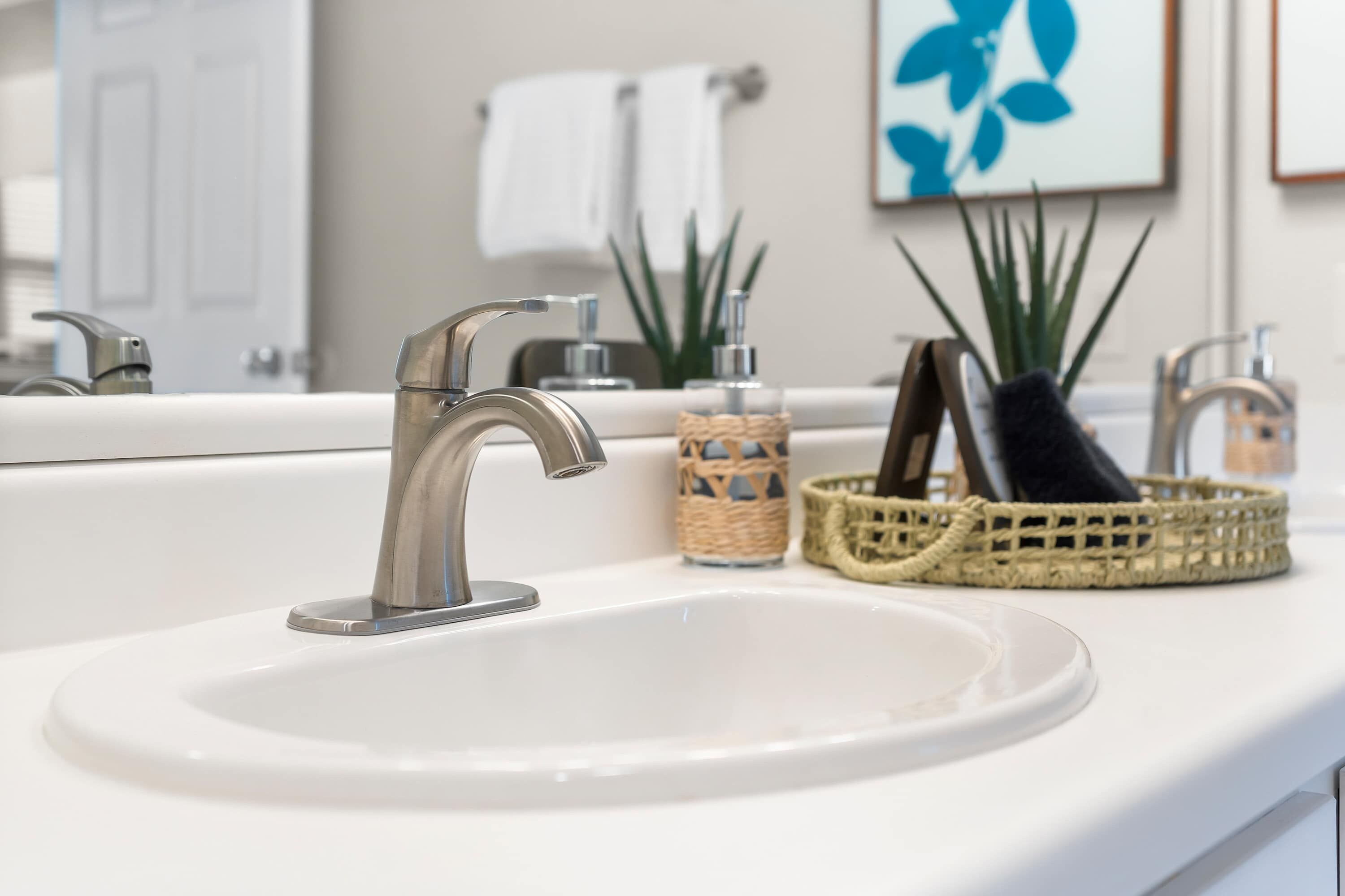 Un lavabo de salle de bain avec un robinet moderne, un comptoir blanc et un miroir au-dessus. On peut voir deux paniers en osier sur le comptoir, l’un contenant une plante et l’autre un objet noir.