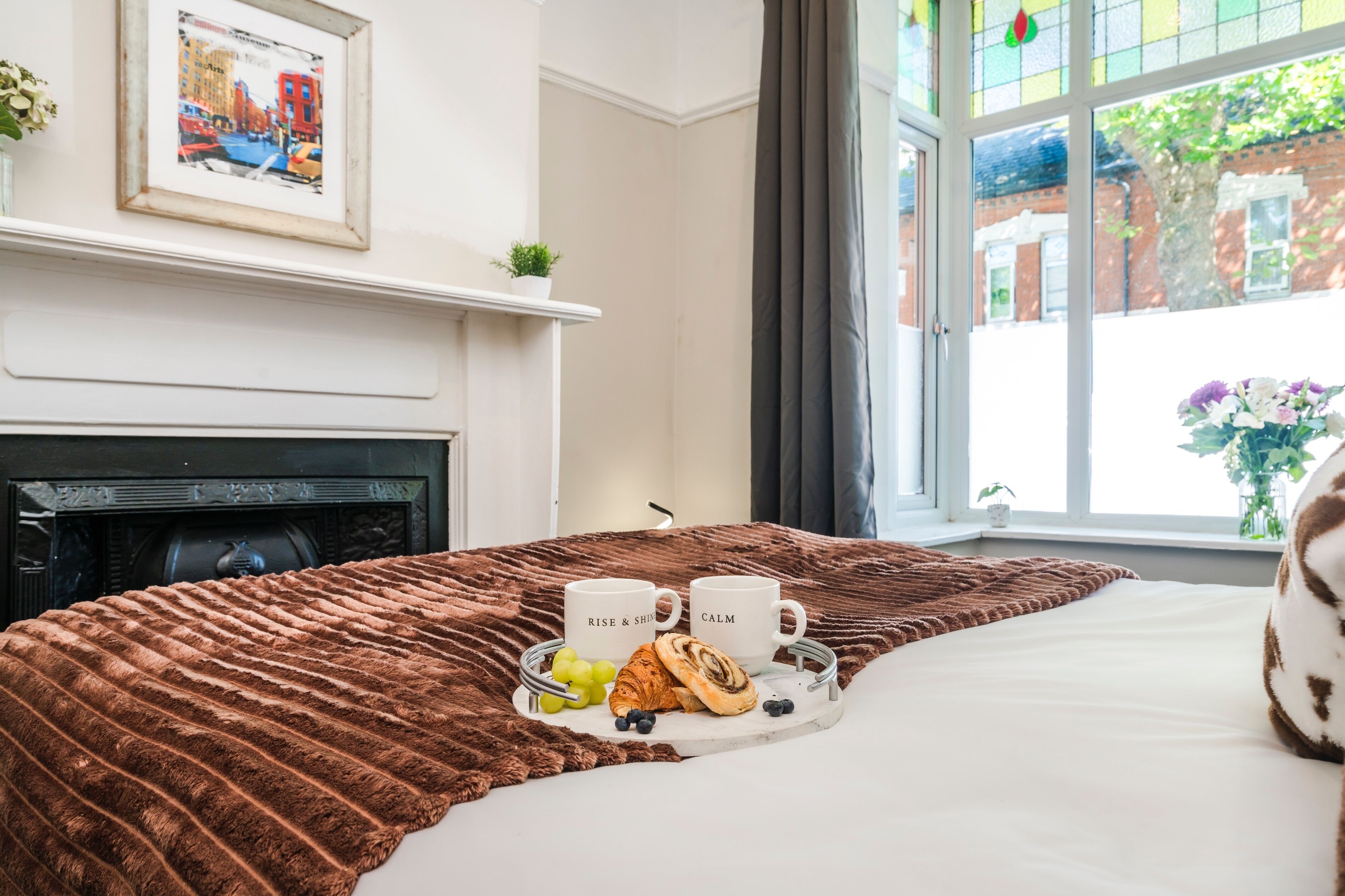 A hotel room with a bed, a tray with croissants, grapes, and two mugs, a fireplace, a framed picture, and a window with a view of buildings and greenery.