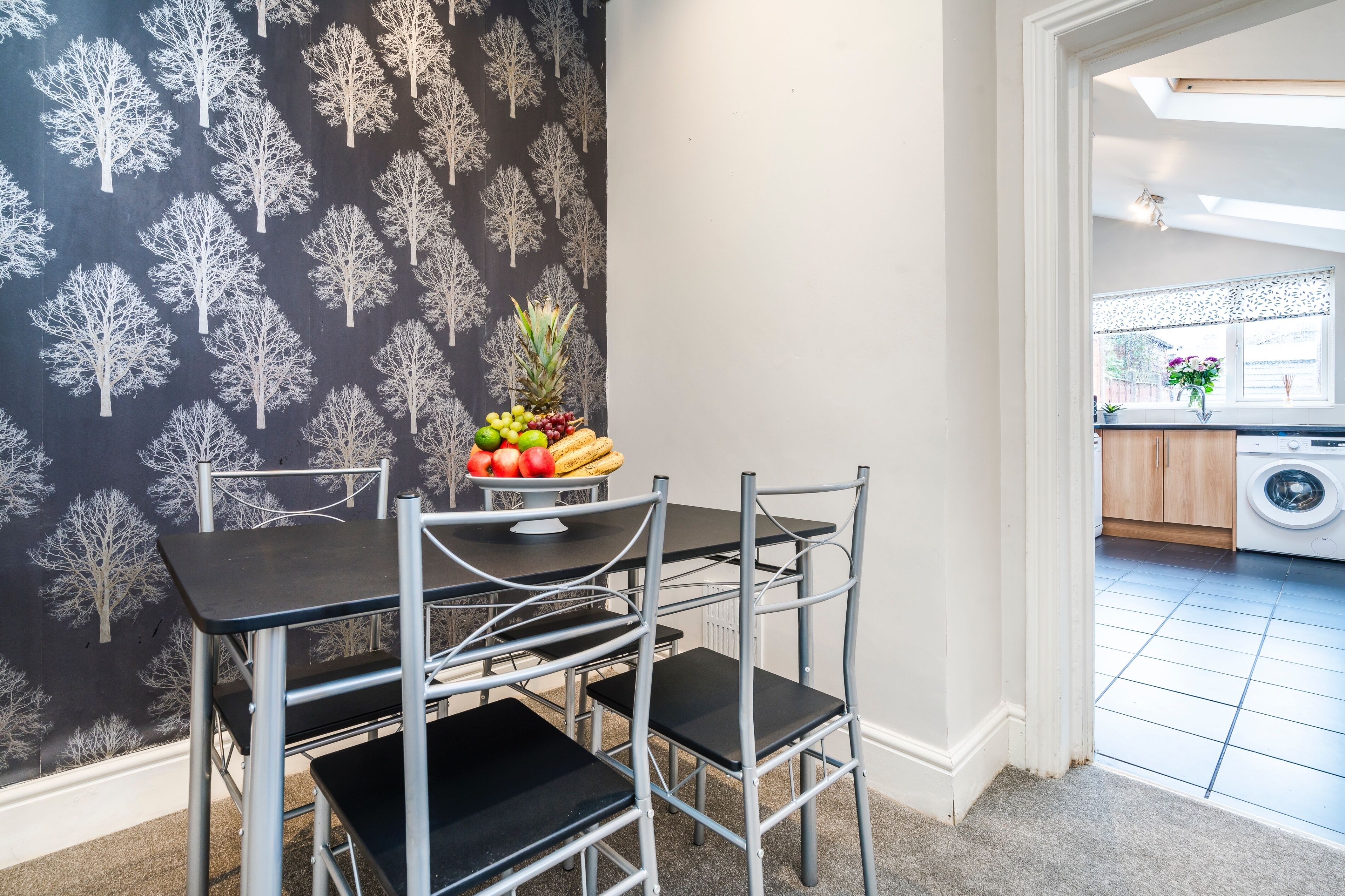 A dining area with a black table and chairs, a fruit bowl on the table, and a kitchen visible in the background.