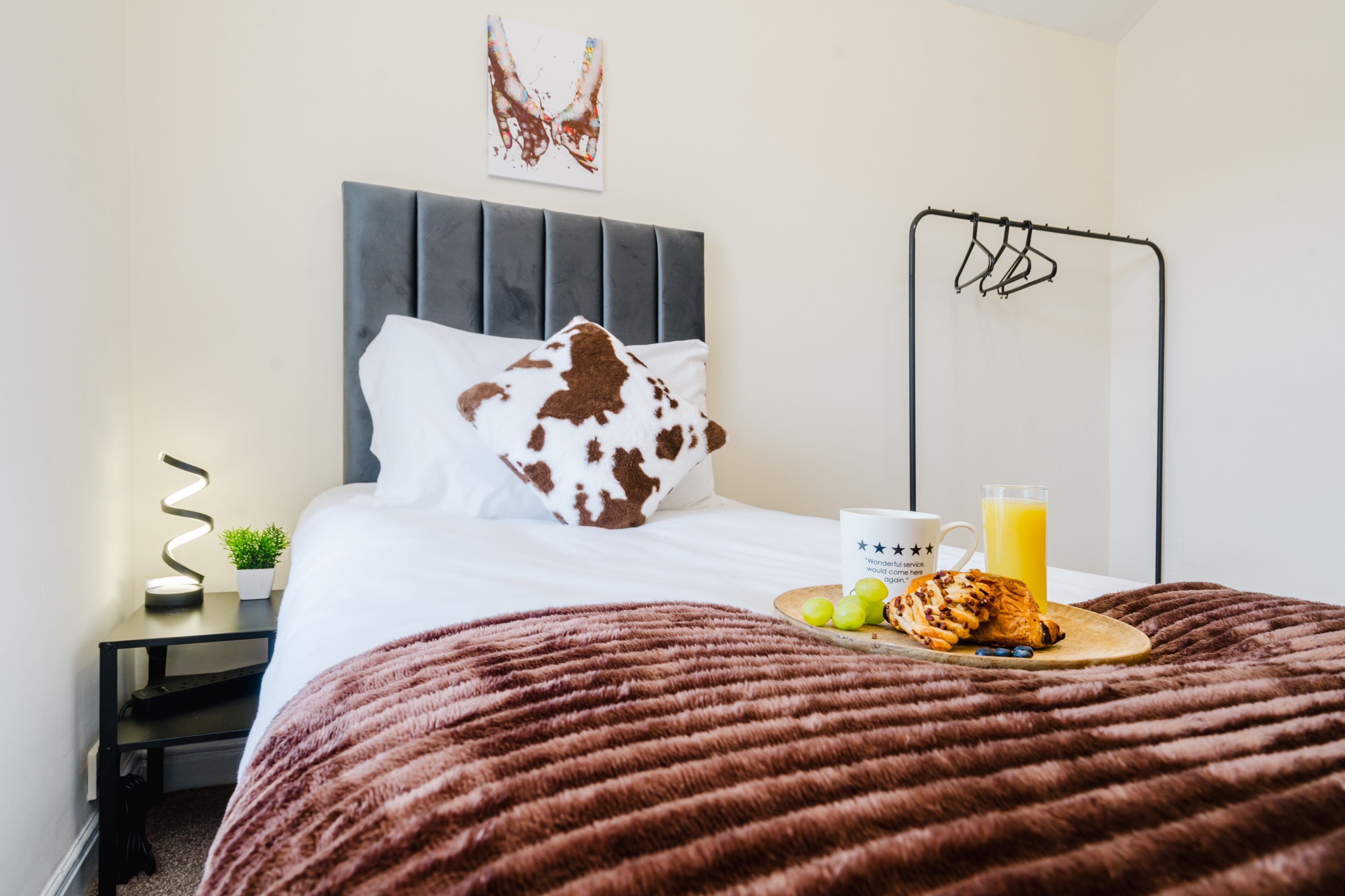 Un lit bien fait, avec un oreiller à motifs marron et blanc, une table de chevet avec une lampe, une assiette de fruits, un croissant et un verre de jus d’orange.