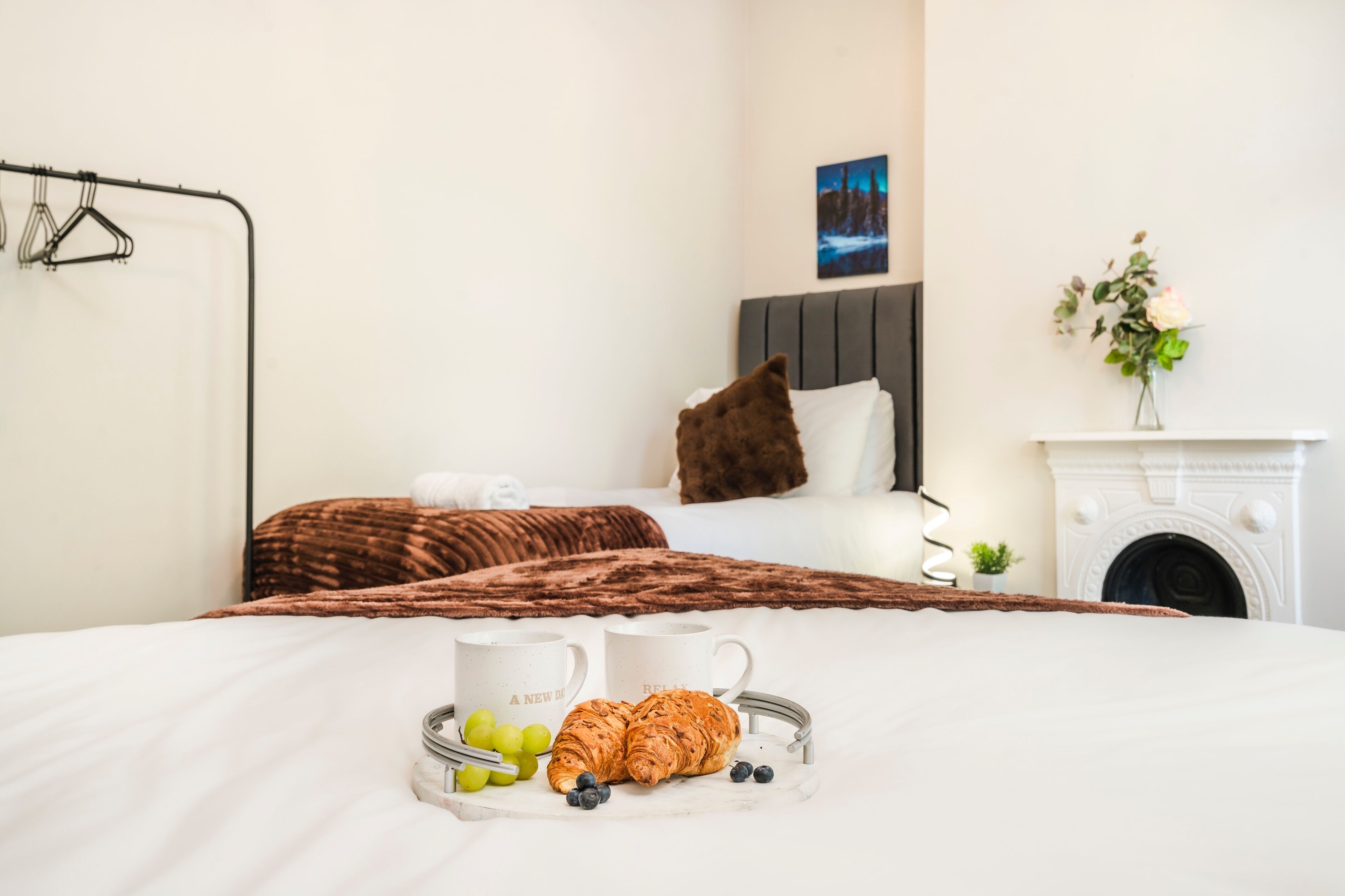 A neatly made bed with a tray of croissants, grapes, and blueberries, a white wall with a painting, and a fireplace.