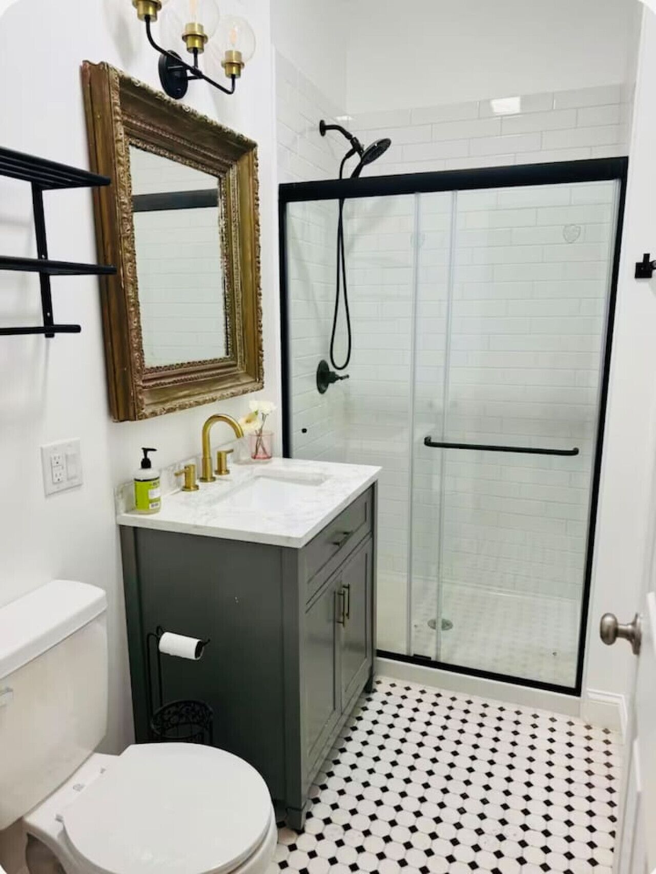 Une salle de bain avec des toilettes blanches, un lavabo avec un robinet doré, un miroir avec un cadre doré et une douche avec une porte noire.