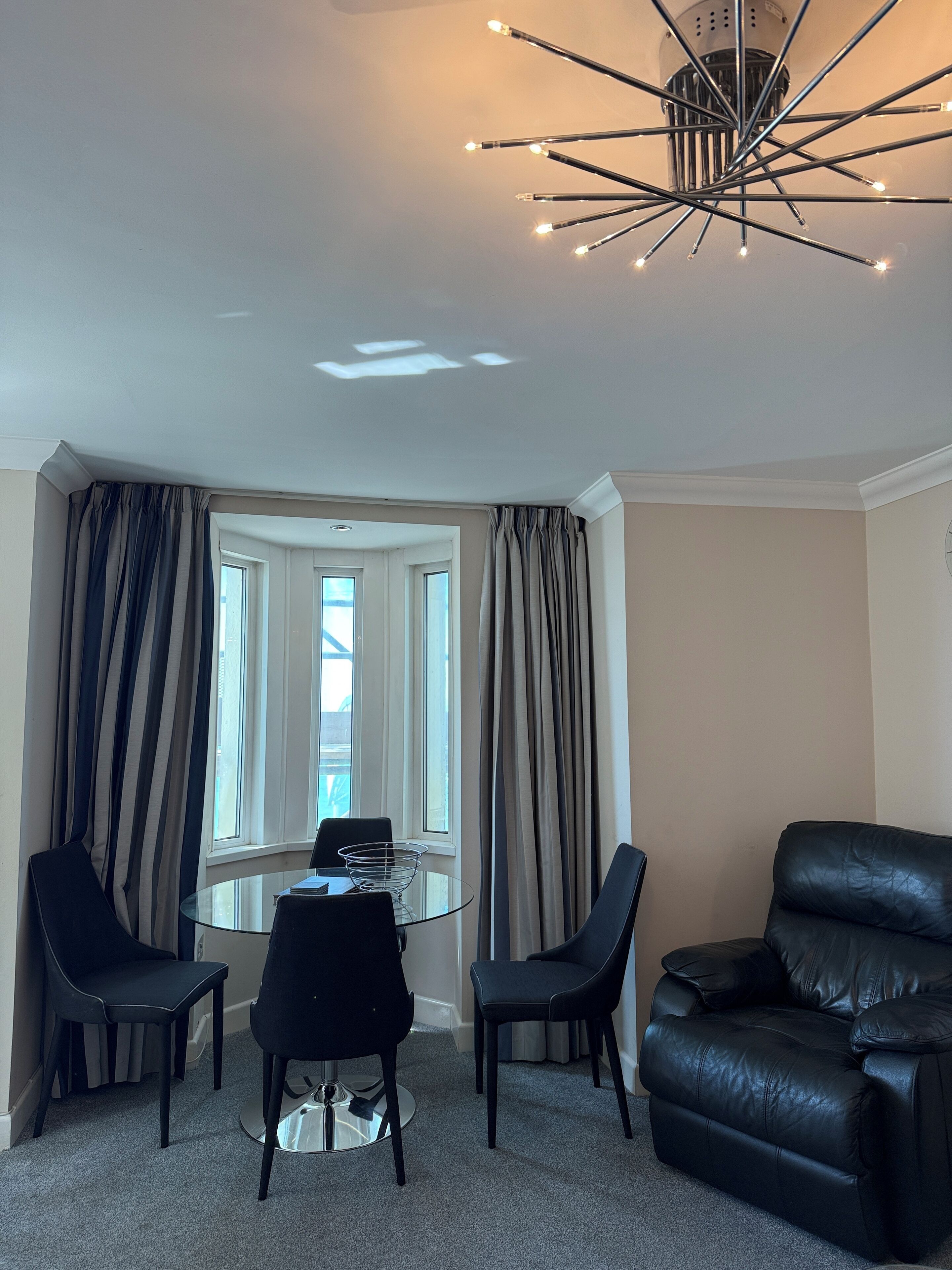 A modern living room with a glass dining table, two black chairs, a black leather sofa, and a unique ceiling light fixture.