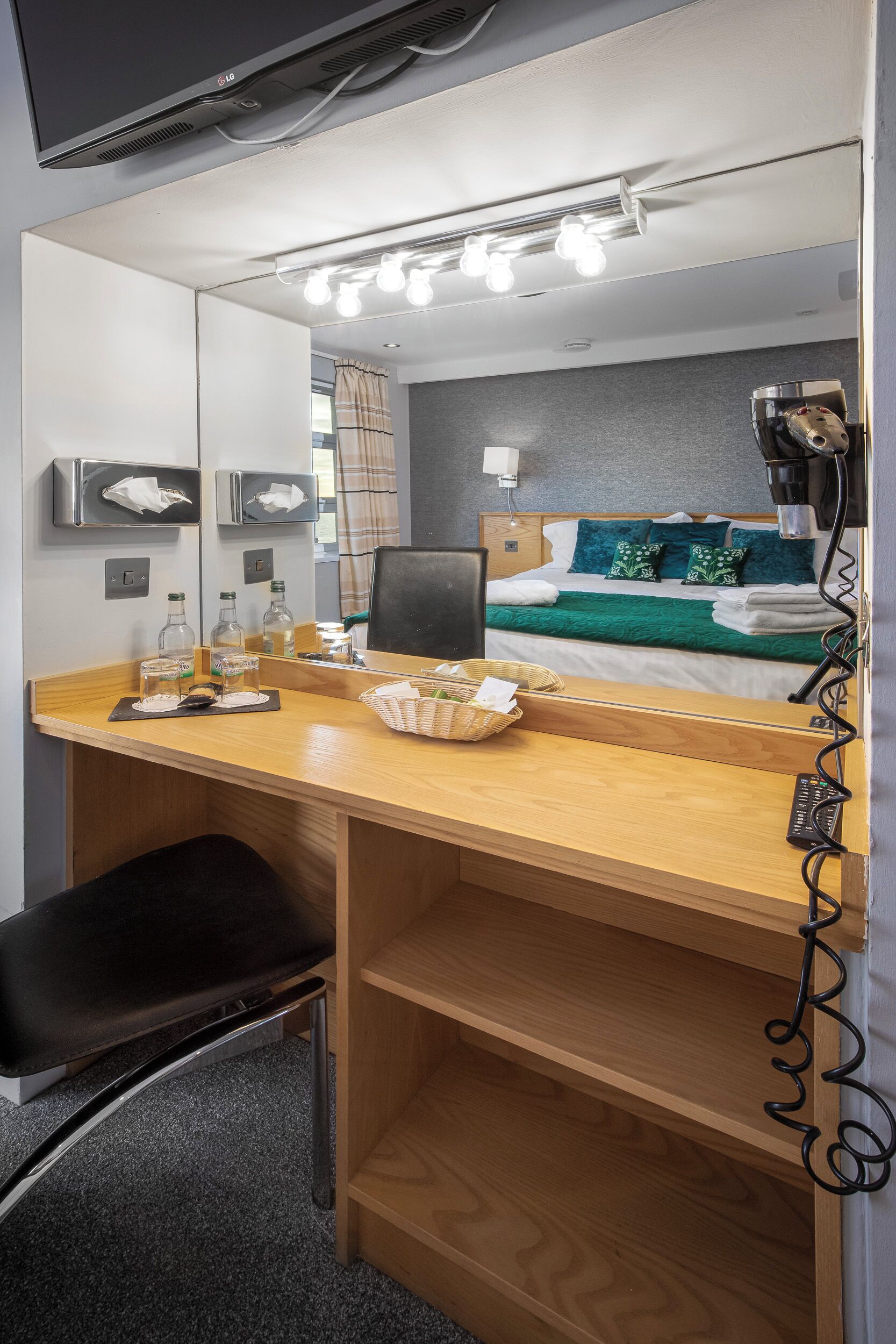 A hotel room with a wooden desk, a black chair, a bed with green and white bedding, a mirror with lights, and a TV mounted on the wall.