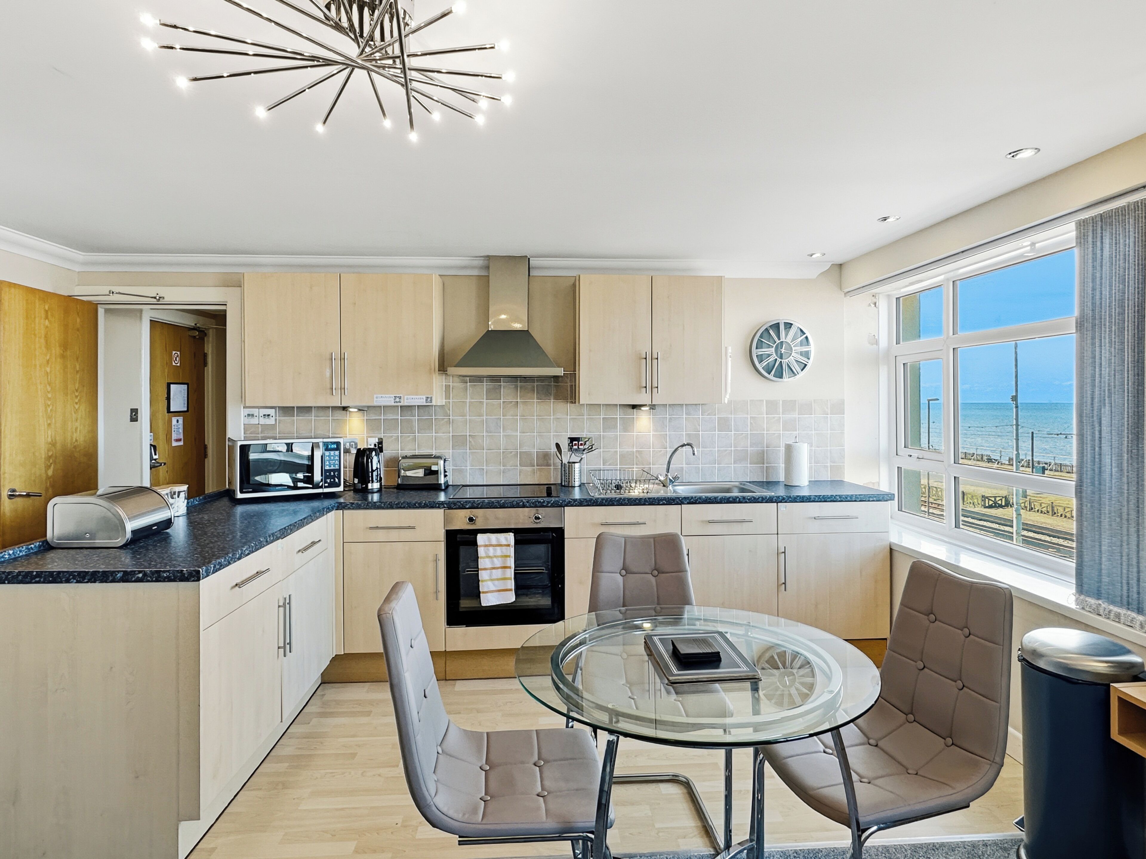 Una cocina moderna con comedor, que cuenta con una mesa de vidrio redonda y sillas acolchadas, horno empotrado y vistas al mar a través de amplios ventanales.