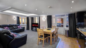 Un salon moderne avec un canapé, une table à manger et des chaises, un téléviseur affichant le logo « NETFLIX », et une vue dégagée grâce à de grandes fenêtres.