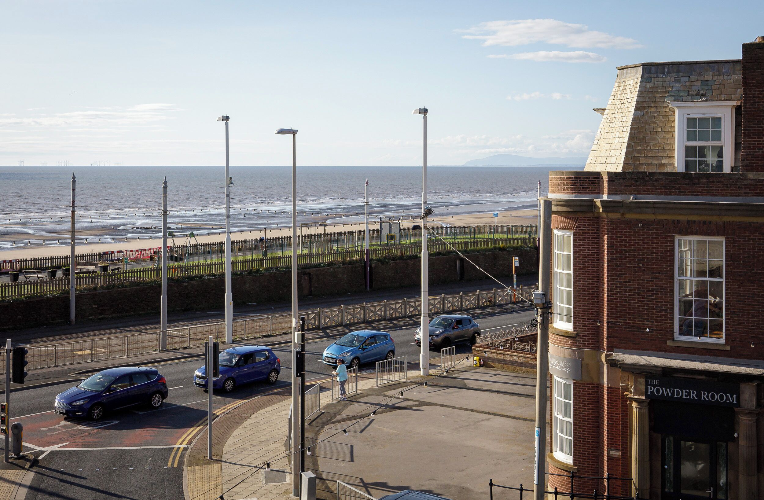 A coastal street with a view of the sea, a building with a sign 'The Powder Room', and parked cars.