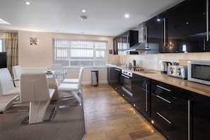 Una cocina moderna con armarios negros, electrodomésticos de acero inoxidable y una encimera de madera. Hay una zona de comedor con sillas blancas y una mesa de vidrio.
