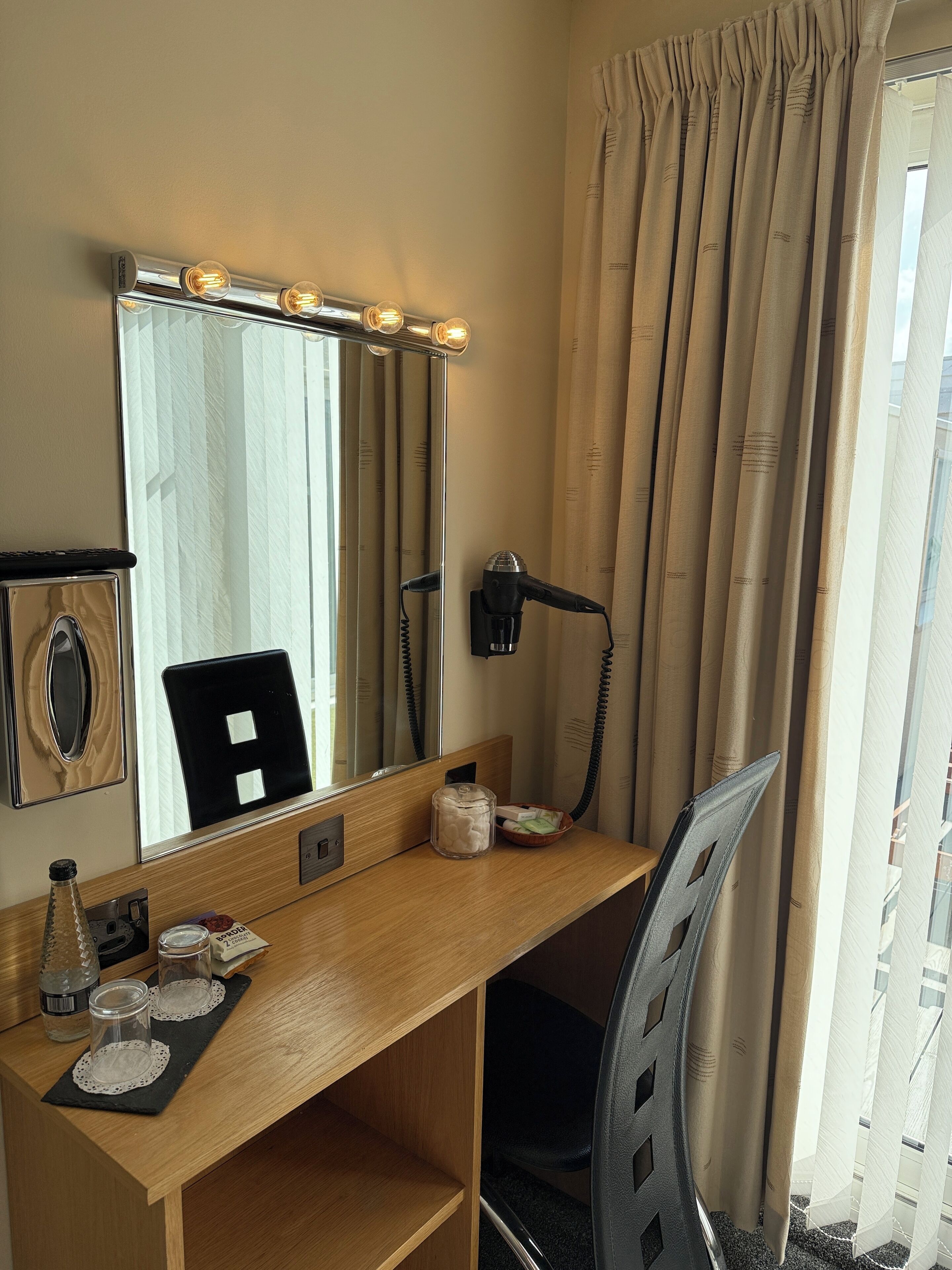 Habitación de hotel con un escritorio de madera, un espejo con iluminación y un secador de pelo, además de una silla.