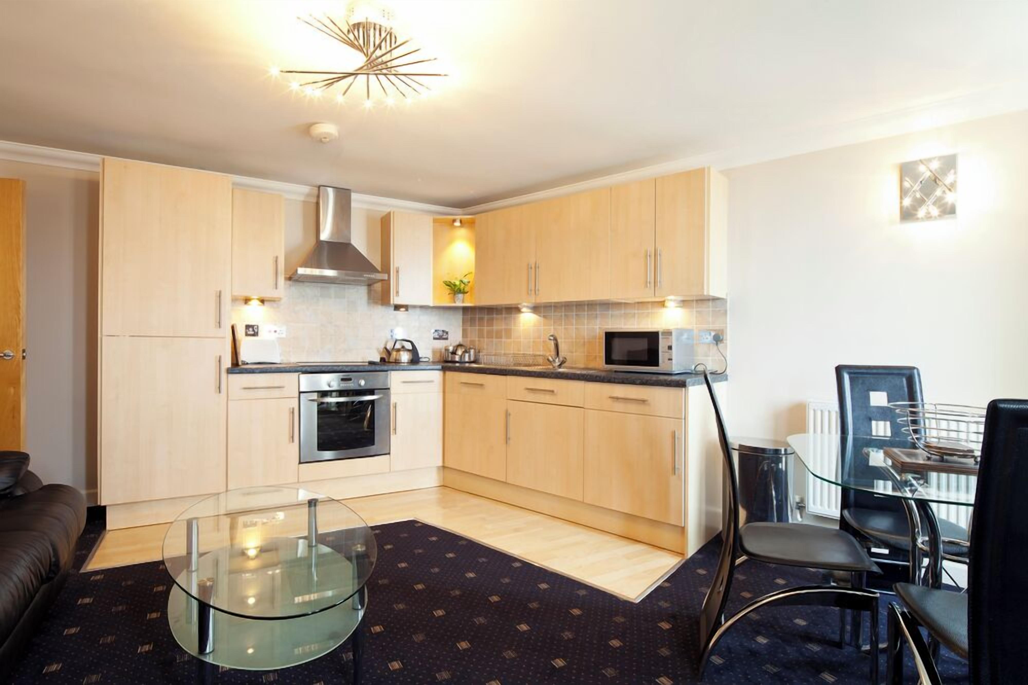 A compact kitchen with light wood cabinets, stainless steel appliances, a glass table, and a dining area with chairs.