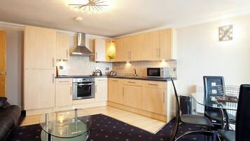 A compact kitchen with light wood cabinets, stainless steel appliances, a glass table, and a dining area with chairs.