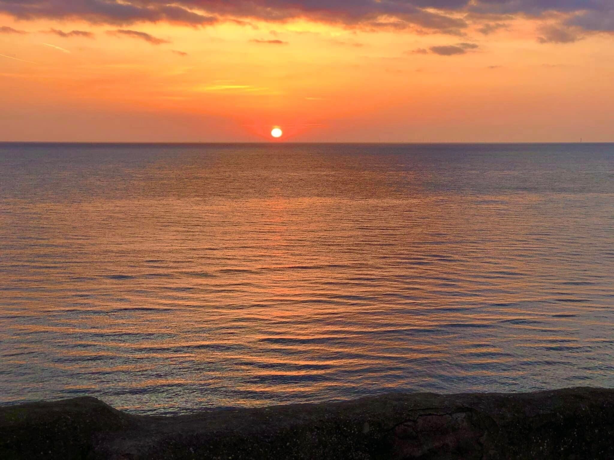 A sunset over a calm sea with a rocky shoreline in the foreground.