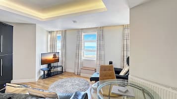 A modern living room with a flat-screen TV, a glass coffee table, a sofa, and a view of the sea through large windows.