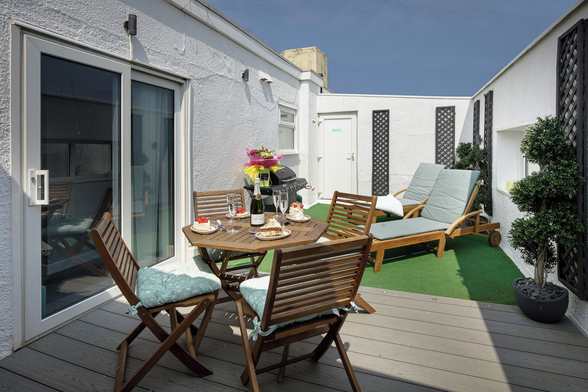 Une terrasse avec une table dressée pour un repas, des sièges d’extérieur et une plante en pot.