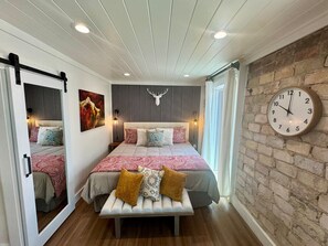 A bedroom with a bed, a bench, a clock, a mirror, and a wall with a brick pattern. - Luxury Suite in the heart of downtown Blue Ridge - Suite 102 (Blue Ridge)