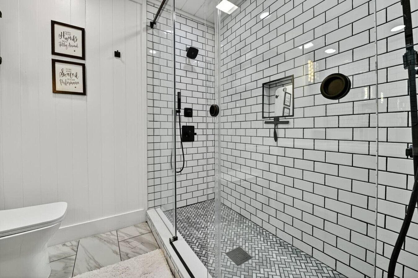 Une salle de bain avec des carreaux de céramique blancs rectangulaires, une douche en verre, des toilettes et deux citations inspirantes encadrées au mur.