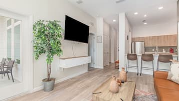A modern living room with a flat-screen TV, a dining area with a wooden table, and a kitchen with bar stools.