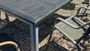 An outdoor dining area with a metal table and wicker chairs, set on a gravel surface with a pond and greenery in the background.