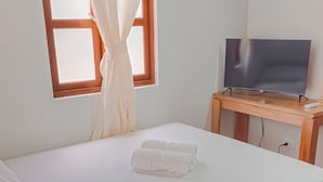 A single bed with white linens and two towels, a wooden nightstand with a television, and a window with curtains. - Hotel Villa Santa Maria  (Santa Fe de Antioquia)
