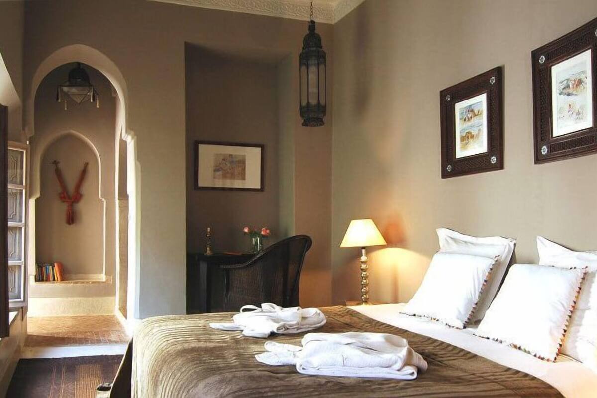 A hotel room with a bed, a desk, a chair, a lamp, and two framed pictures on the wall.