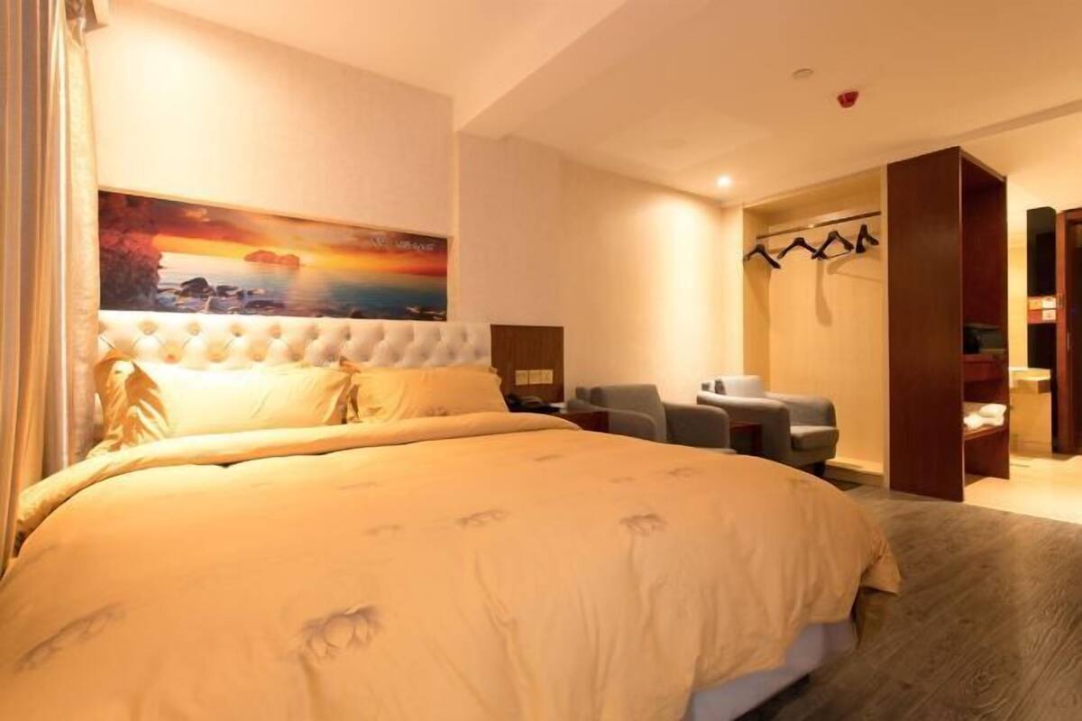 A hotel room with a large bed, a seating area with a chair, a painting on the wall, and a coat rack.
