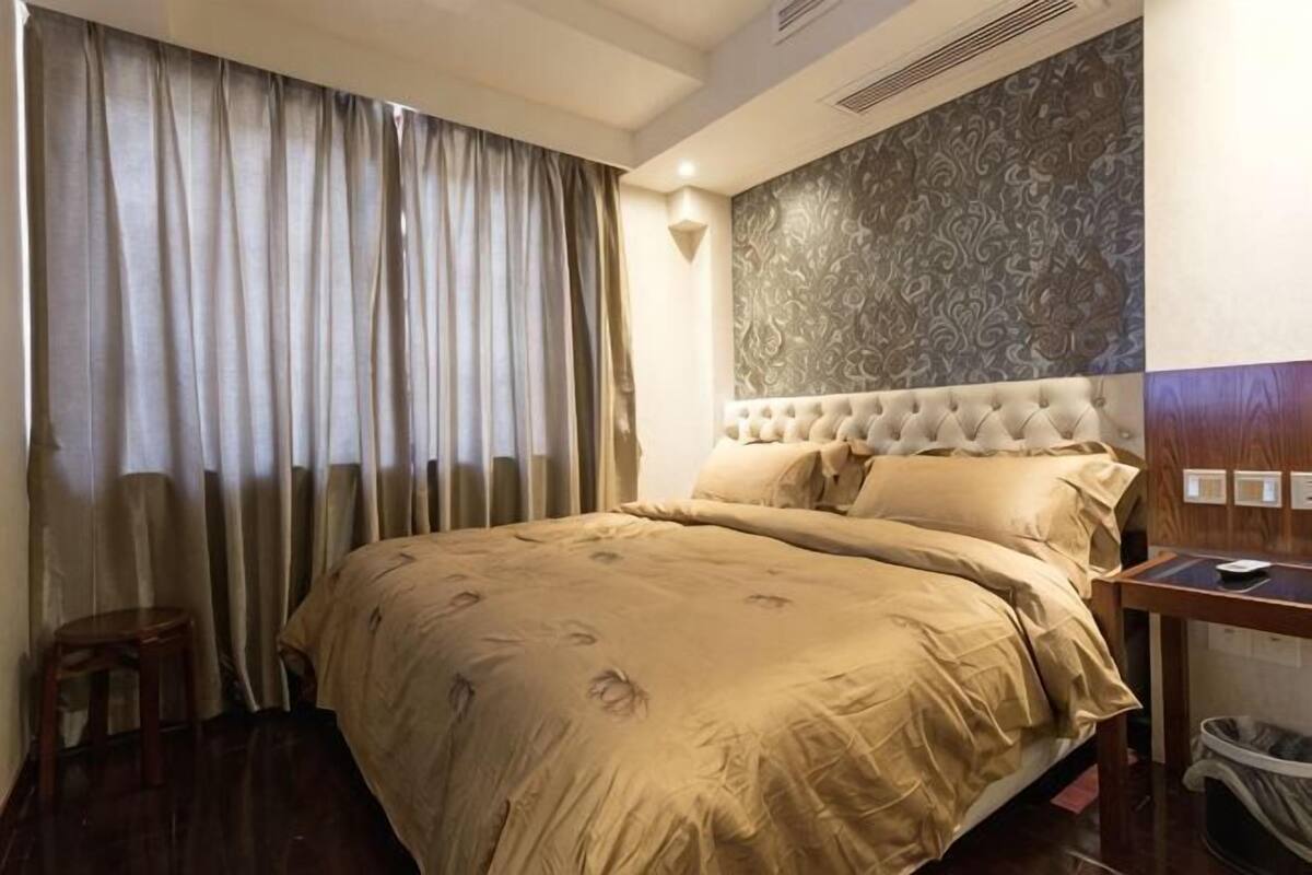 A hotel room with a large bed, a headboard with a patterned design, a small wooden stool, and a wall-mounted light fixture.