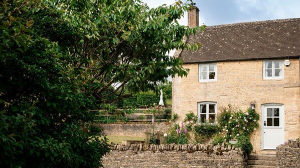 A stone house with a brick roof, white doors, and windows, surrounded by lush greenery and a stone wall.