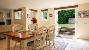 A dining area with a wooden table, white chairs, and a vase with flowers. There is a window with a view of greenery outside. - Pickett Cottage - Pet friendly near Northleach (Northleach)