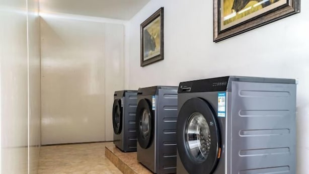A row of three front-loading washing machines in a clean, tiled laundry room with a plain white wall and a framed picture above the machines. - Jiuyuan Hotel (Jiuzhaigou)