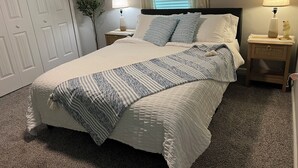 Een opgemaakt bed met een gestreept kleed, twee bedlampen, een nachtkastje, een kamerplant en een raam met jaloezieën.