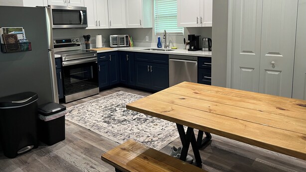 Een moderne keuken met witte kasten, roestvrijstalen apparatuur, een houten eettafel en een vloerkleed met een patroon.